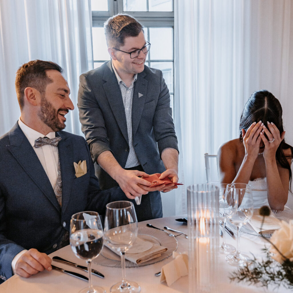 Bräutigam lacht, die Braut schlägt staunend die Hände vors Gesicht – Tischzauberei auf einer Hochzeit